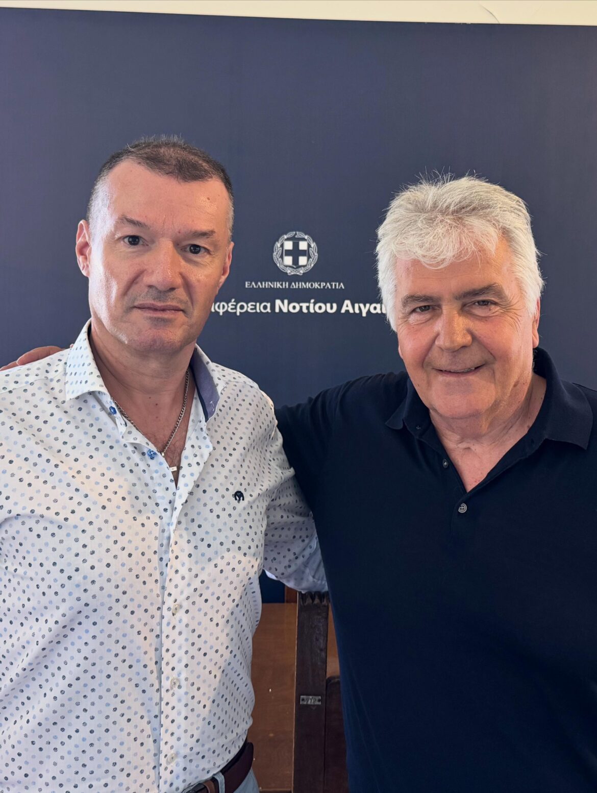 Two men posing together in front of a dark blue backdrop with a Greek government seal and Greek text.