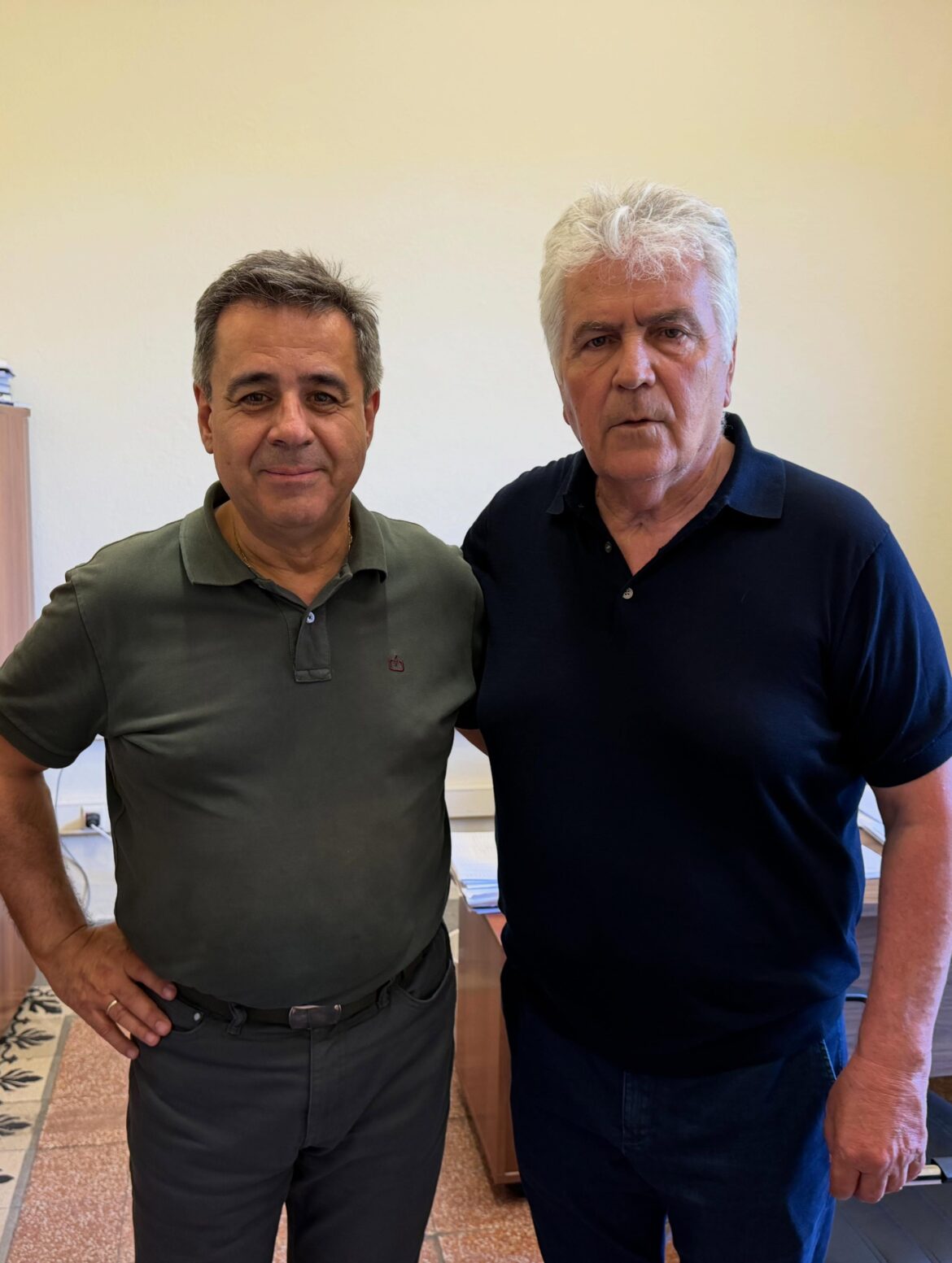 Two middle-aged men standing side by side indoors, facing the camera in casual polo shirts (green and navy).