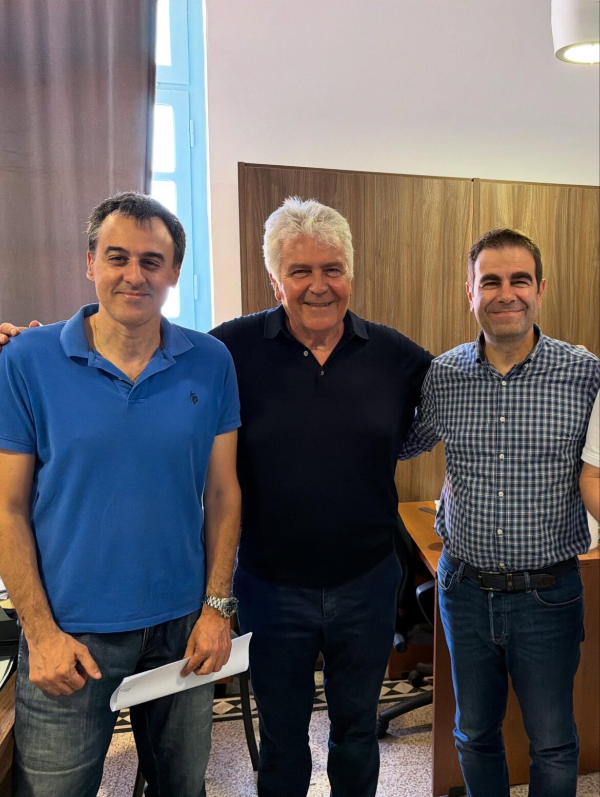 Three men stand together in an office, smiling at the camera with arms around each other.
