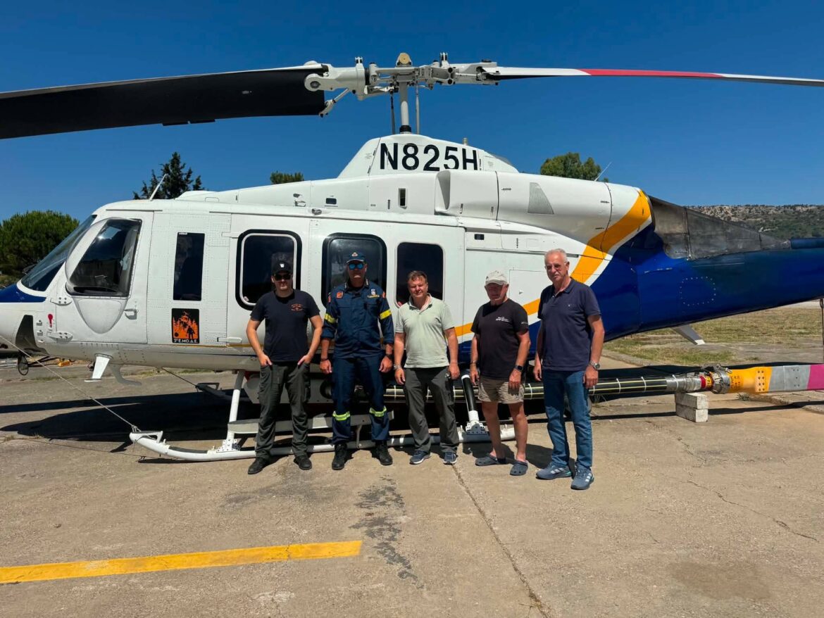 A group of five men posing in front of a white helicopter on a tarmac under a clear blue sky.
