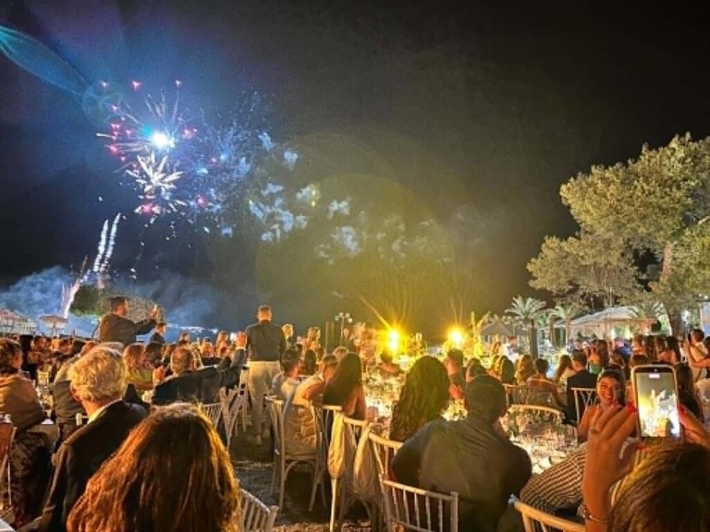 Crowd seated on chairs in a courtyard watching colorful fireworks explode in the night sky in a festive outdoor scene.