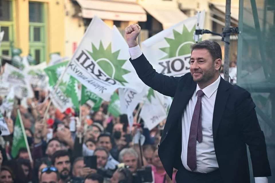 Man in a suit raising his fist on a stage at a political rally, with a cheering crowd and green-and-white flags in the background.