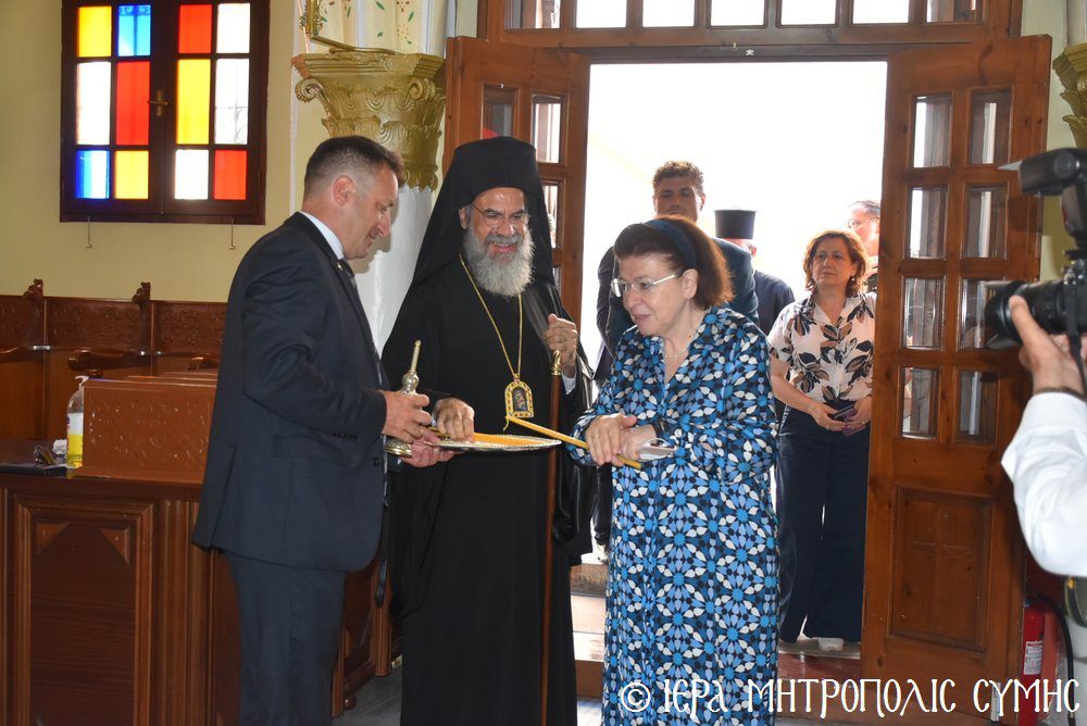 Orthodox priest in black robes mediates a ceremonial exchange as a man in a suit and a woman in a blue patterned dress hand over a document inside a church.