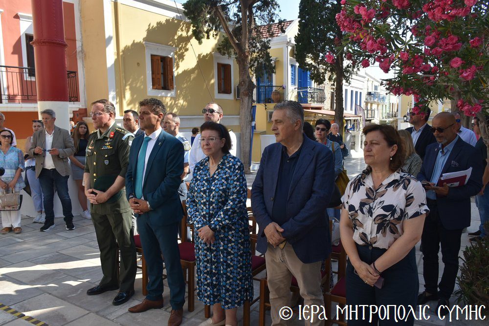 Group of formally dressed people standing in a sunlit town square during a ceremony; includes a man in a military uniform and others in suits.