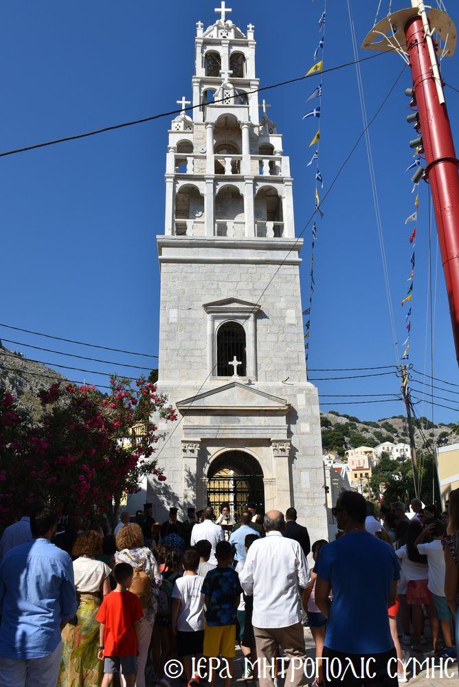 Stone church tower with multiple arches rises above a crowd gathered at its entrance on a sunny day, flag lines and flowering bushes nearby.