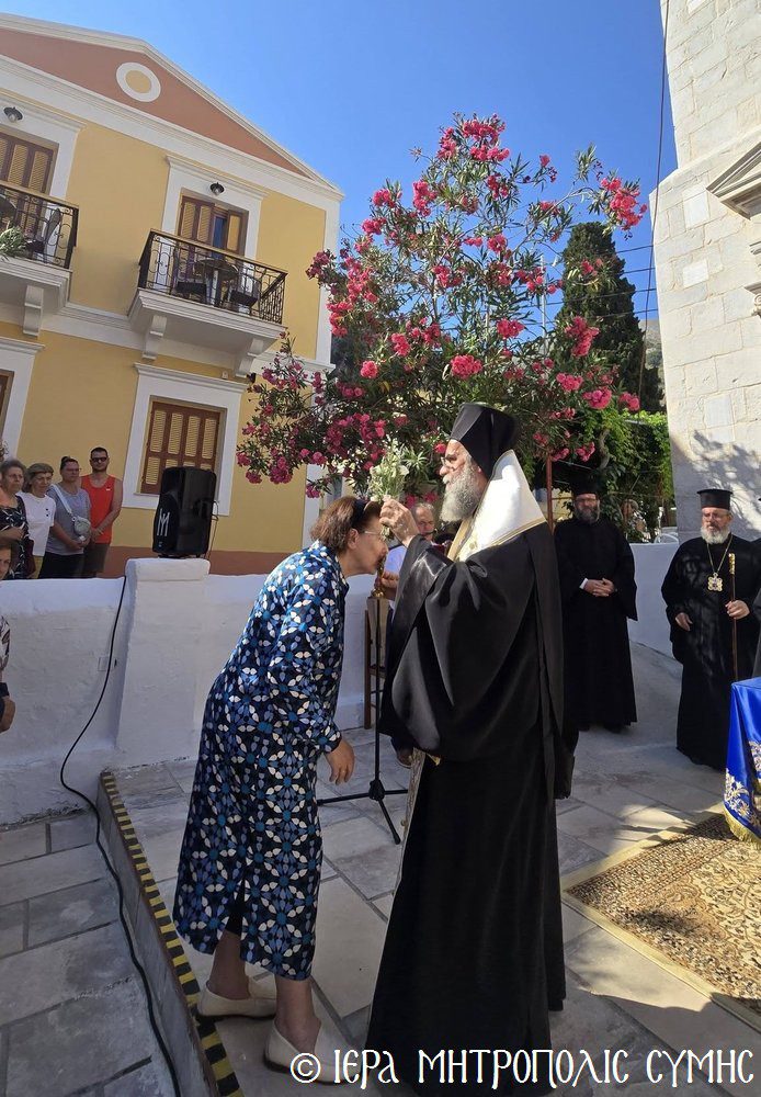 A Greek Orthodox archbishop in black robes blessing a woman who kisses his hand, outdoors beside a yellow building and pink flowering tree with onlookers nearby.