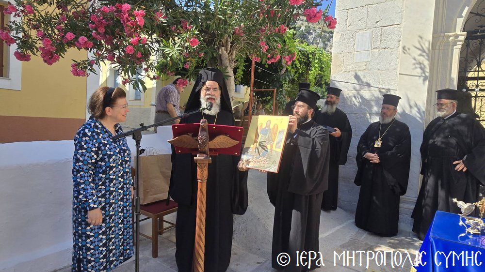 Orthodox ceremony outdoors with a priest at a wooden podium speaking into a microphone as another man holds a religious icon; a woman in a blue patterned dress stands nearby, with several clergymen in black robes watching under pink flowering vines by a stone building.