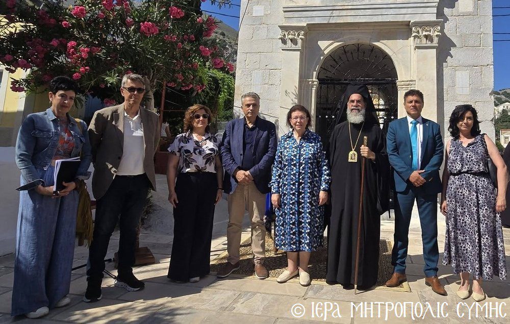 Ten people pose for a group photo outside a stone church, led by an Orthodox priest in black robes with a gold cross.