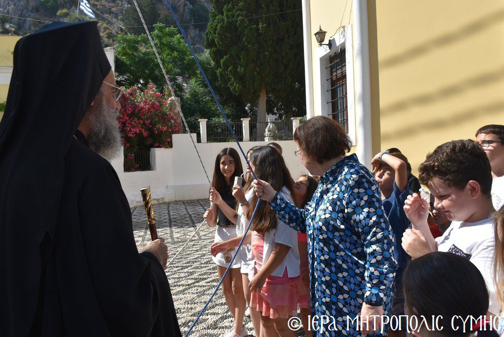 Orthodox priest in black robes speaking to a group of children and an adult woman outside a yellow building with a Greek flag in the background.