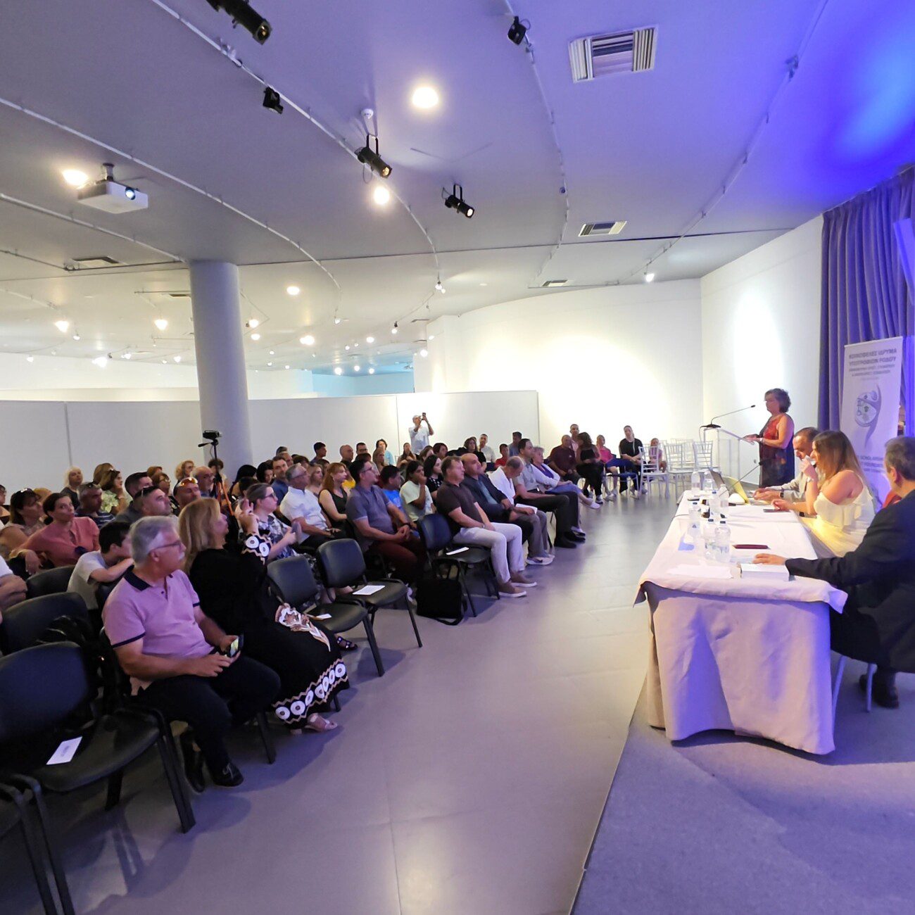 Panel discussion in a bright conference room with a speaker at a podium and a row of panelists seated at a long table facing an audience of about 50 people.