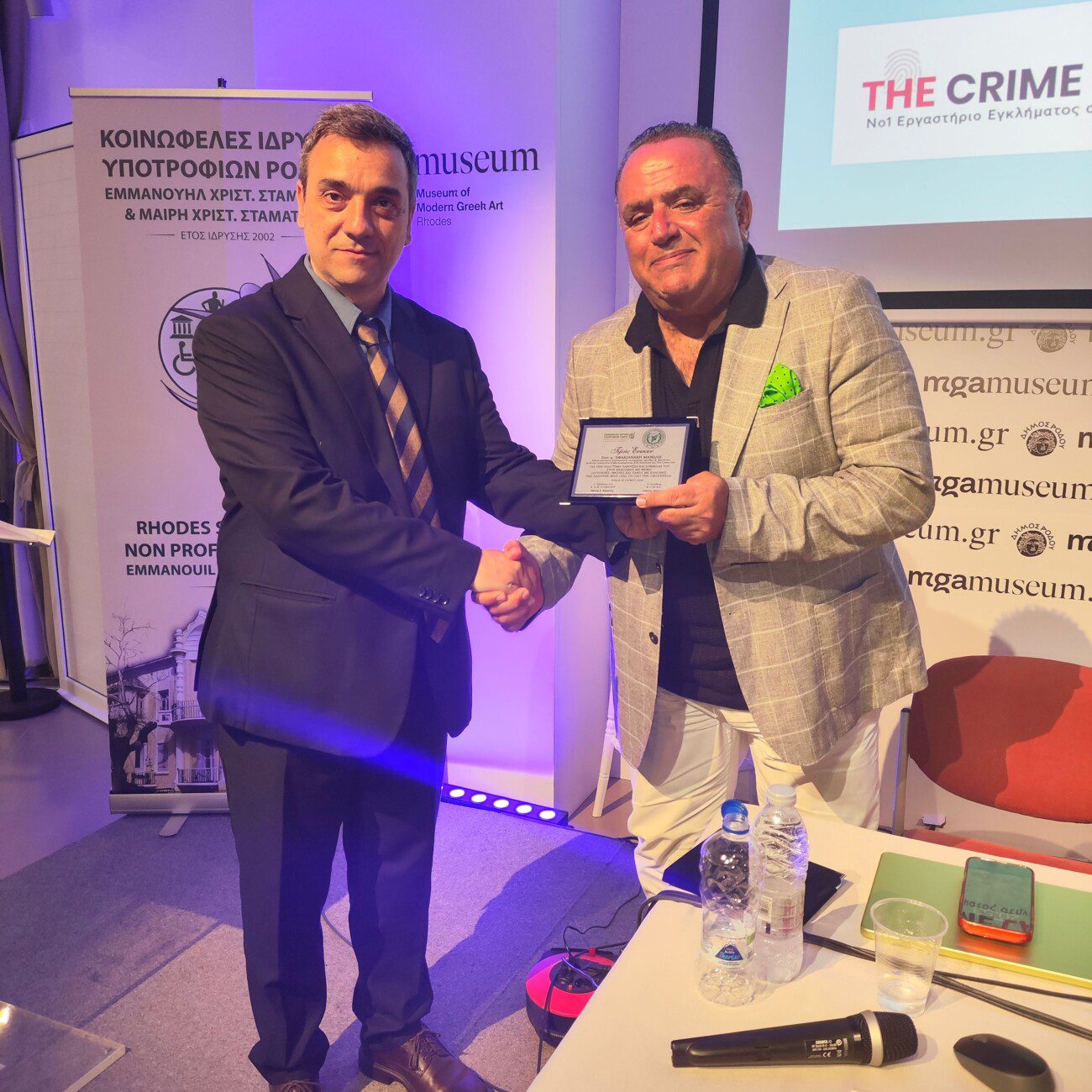 Two men in suits shake hands as one receives a plaque at an awards ceremony, with banners in the background.