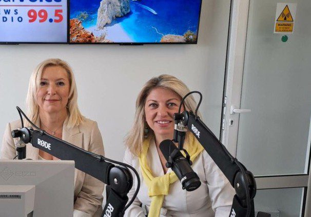 Two women sit in a radio studio speaking into Rode microphones, with a computer monitor and screen behind them.