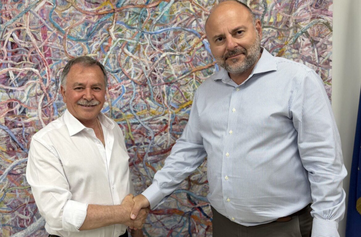 Two men shake hands in front of a large colorful abstract painting, one wearing a white shirt and the other a light blue striped shirt.