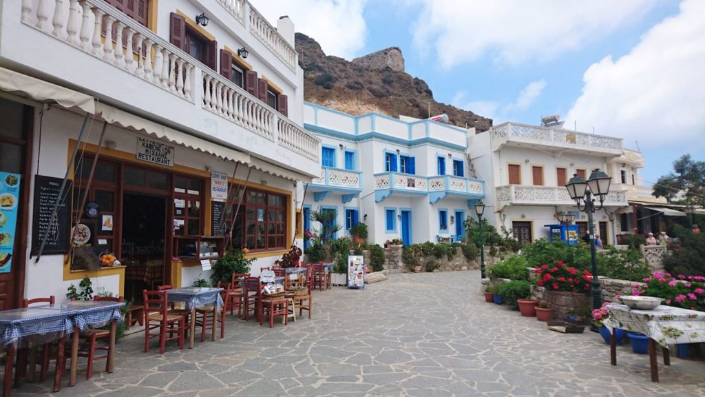 Sunny Mediterranean street with white and blue buildings, outdoor cafe tables along a stone-paved walkway and potted plants.