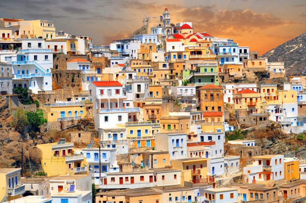 Colorful hillside village with white, blue, yellow houses stacked on terraces under a warm sunset sky.