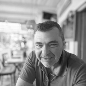 Smiling man in a polo shirt sits in a bright cafe, photographed in black and white.