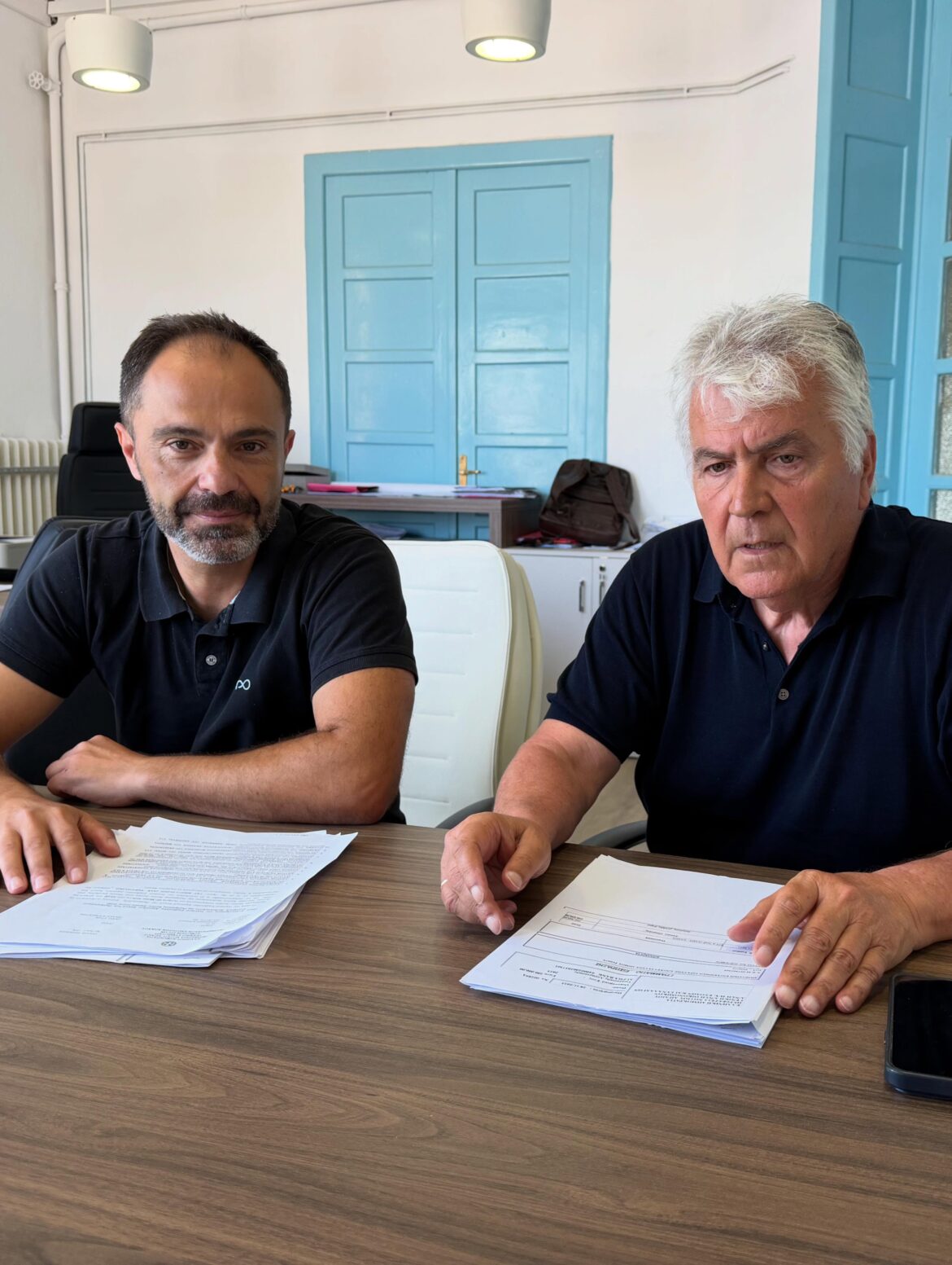 Two men in polo shirts sit at a wooden table reviewing and signing papers in a bright office with blue doors in the background.
