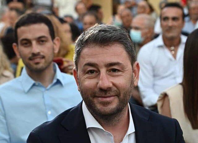 Smiling man in a dark blazer and white shirt sits in front at an outdoor gathering, with a blurred crowd behind him.