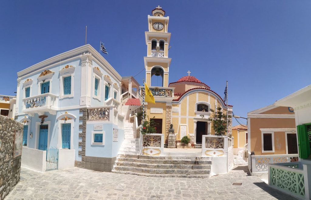 Pastel-blue building with blue doors and balconies beside a small yellow church with a bell tower and red-tiled dome in a sunny Greek village square.