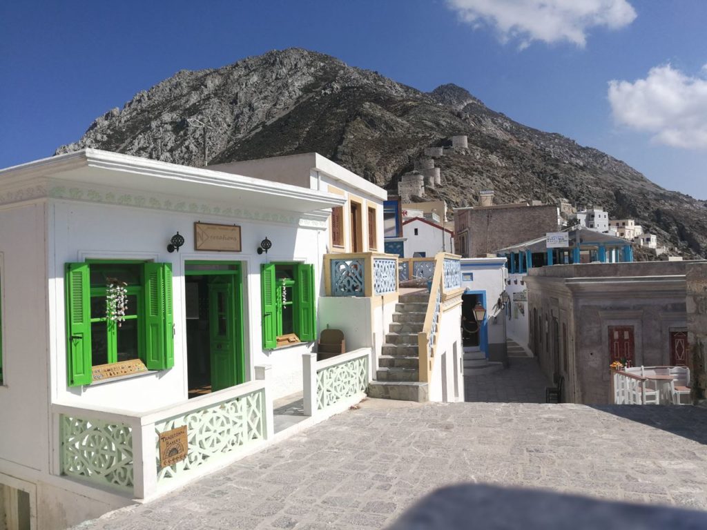 White-washed buildings with bright green shutters along a cobblestone street, against a rocky mountain backdrop on a sunny day.