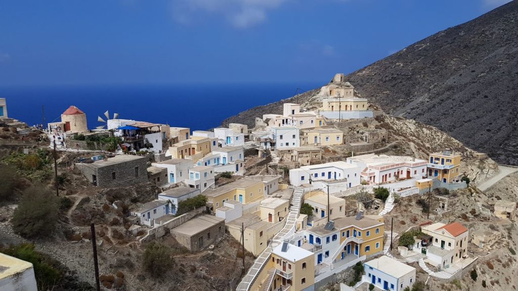 Coastal hillside village with white and pastel houses, stone stairways, and the blue Aegean Sea in the background.