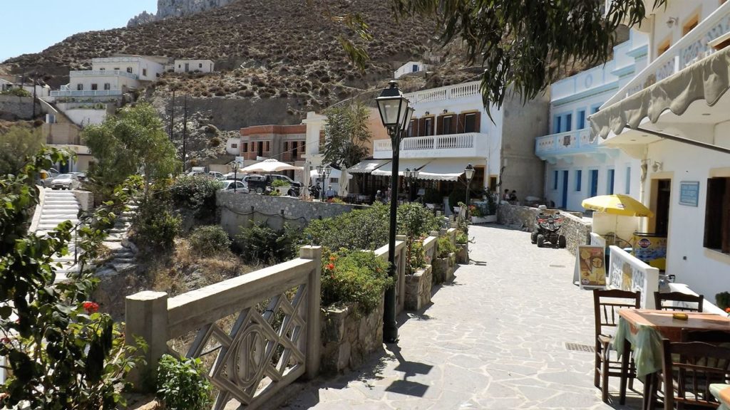 Sunny cobblestone street in a hillside village, with pastel shops, outdoor seating under umbrellas, and a lamppost in the center in bright daylight.