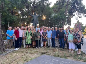 A large group of people posing together in a park, with a statue behind them among trees and greenery.