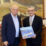 Two men in suits pose in a formal office; one holds a blue book titled 'Aegean Memories'.