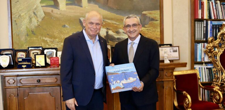 Two men in suits pose in a formal office; one holds a blue book titled 'Aegean Memories'.
