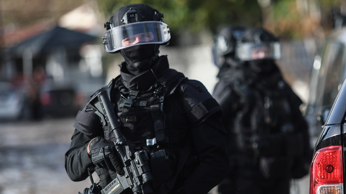 Two heavily armed tactical officers in black gear and clear-face shields stand near a vehicle during a daytime operation.