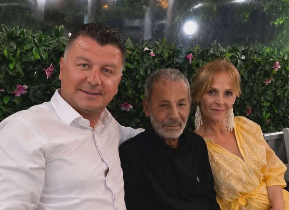Three adults posing for a photo on a bench in a garden with a flowering hedge behind them at night.