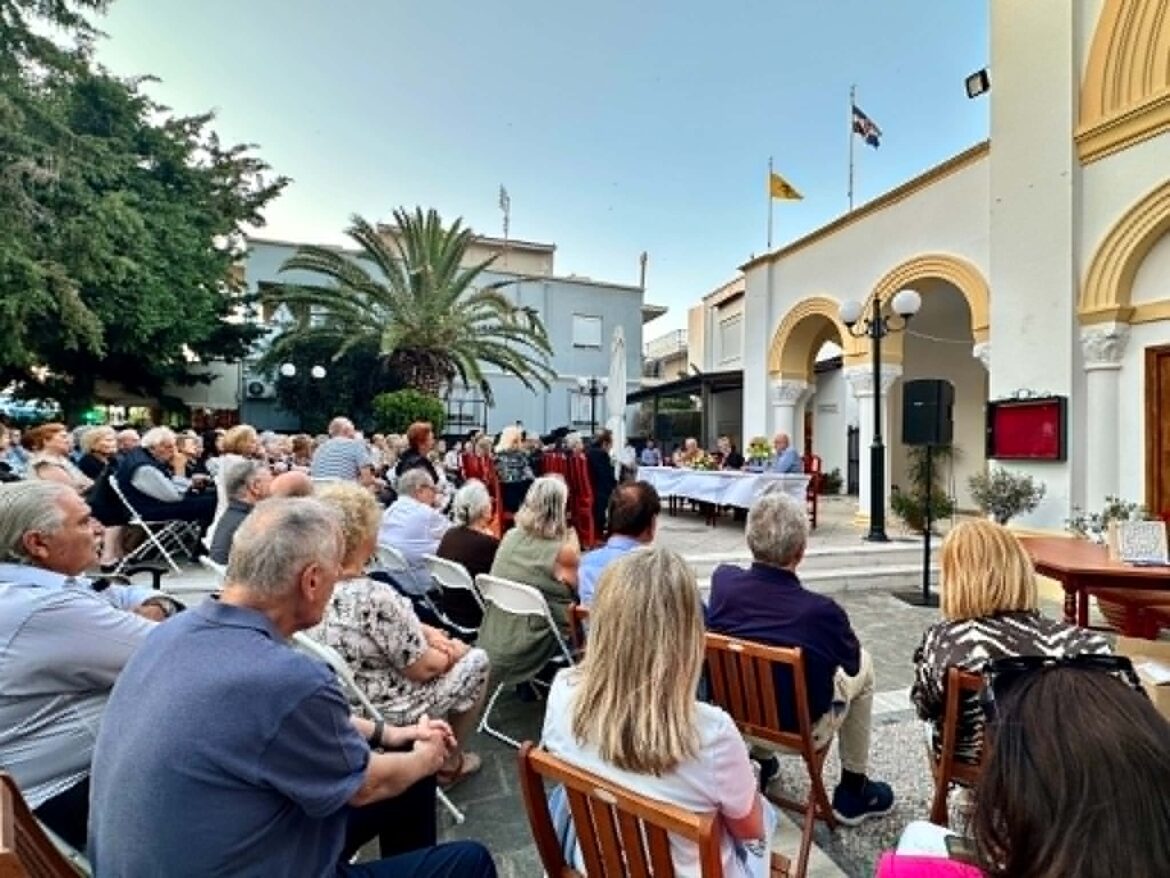 Crowd seated outdoors for a ceremony in a town square with a cream building and flags in the background.