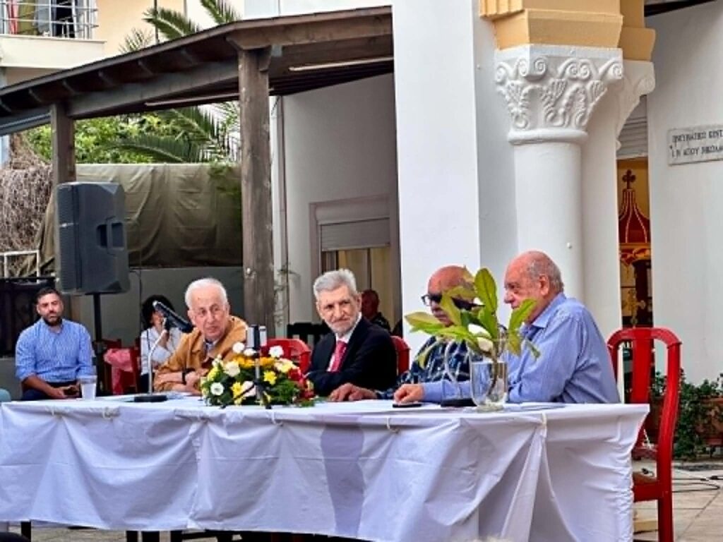 Group of men seated at a long table outdoors, with a floral centerpiece in front and a white tablecloth.