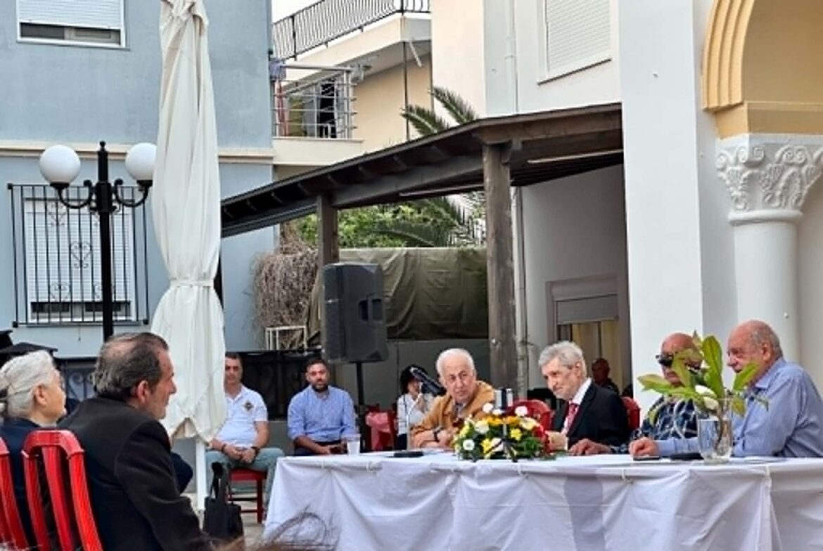 Group of people seated around a long table in an outdoor courtyard, floral centerpiece, light-colored buildings in the background.