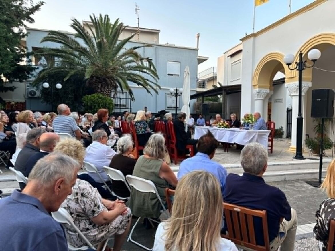 Outdoor ceremony or gathering with many attendees seated in rows, facing a long table covered in white cloth in a sunlit courtyard.