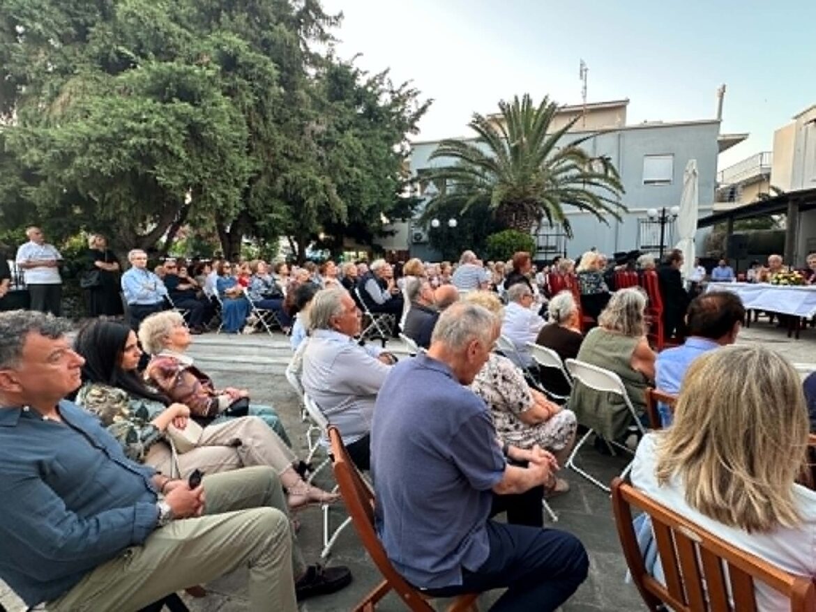 Large crowd seated outdoors for an event, with trees and buildings in the background.