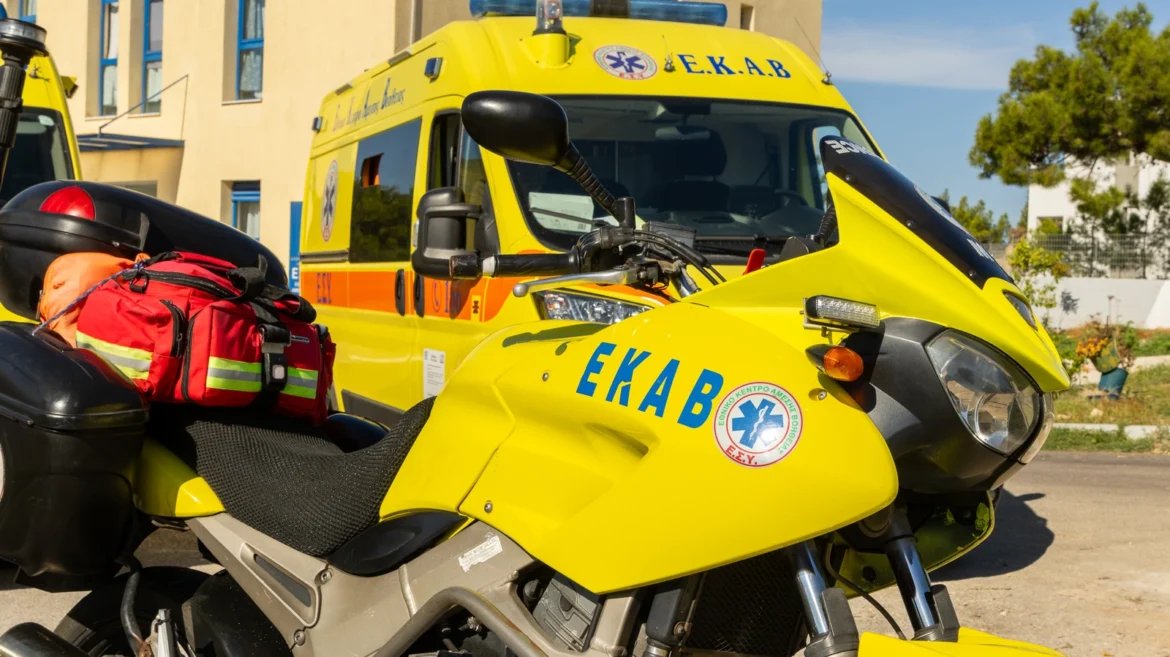 Emergency medical services motorcycle in the foreground with red kit bags, and a yellow ambulance labeled E.K.A.B. in the background.",