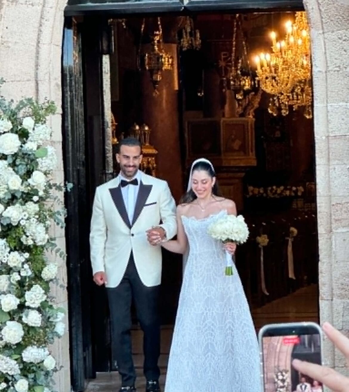 Bride and groom walk out of a church doorway, smiling; she holds a bouquet in a light dress, he in a white suit jacket with a bow tie.