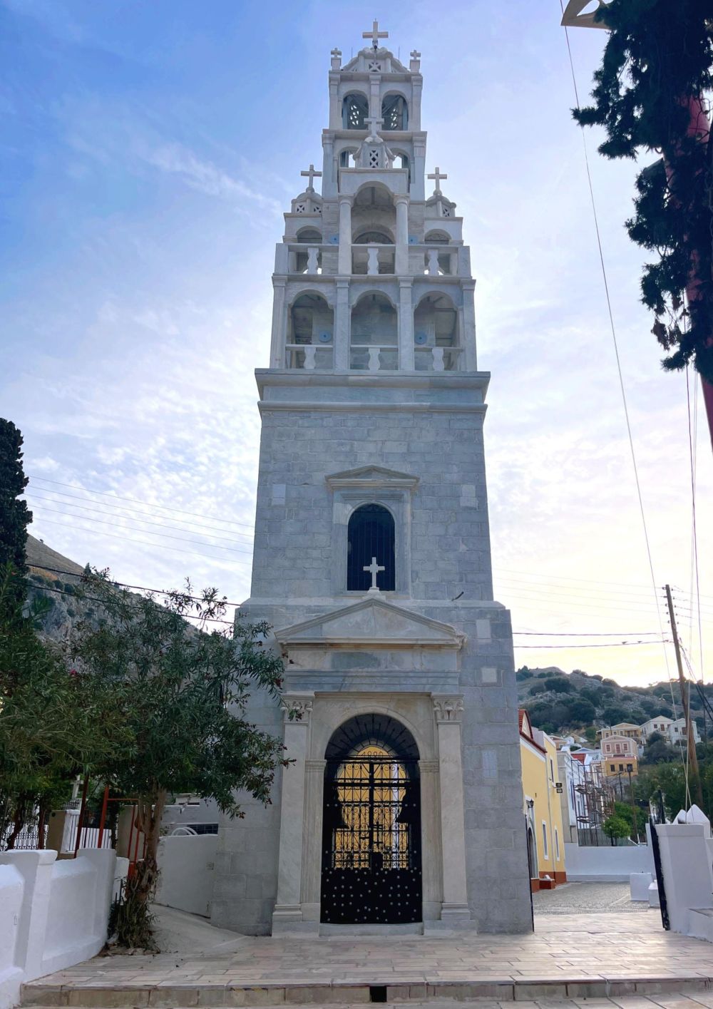 Stone church bell tower with multiple arches and crosses rises against a blue sky; iron-gated entrance at base.
