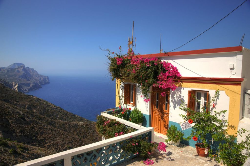 Coastal house with vibrant pink bougainvillea, white and yellow walls, and a view of the blue sea and cliffs beyond the terrace rail.