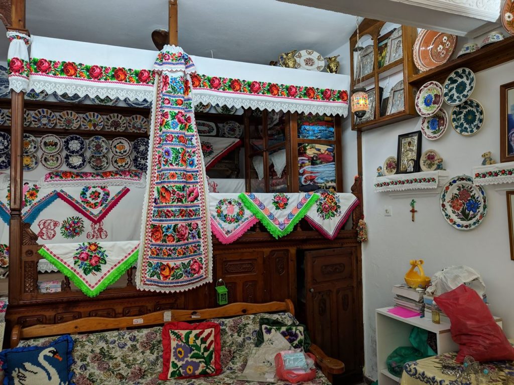 A cozy room filled with colorful folk-art textiles: embroidered table runners and wall hangings draped over a wooden cabinet, with decorative plates on the wall and shelves, and cushions on a floral sofa.