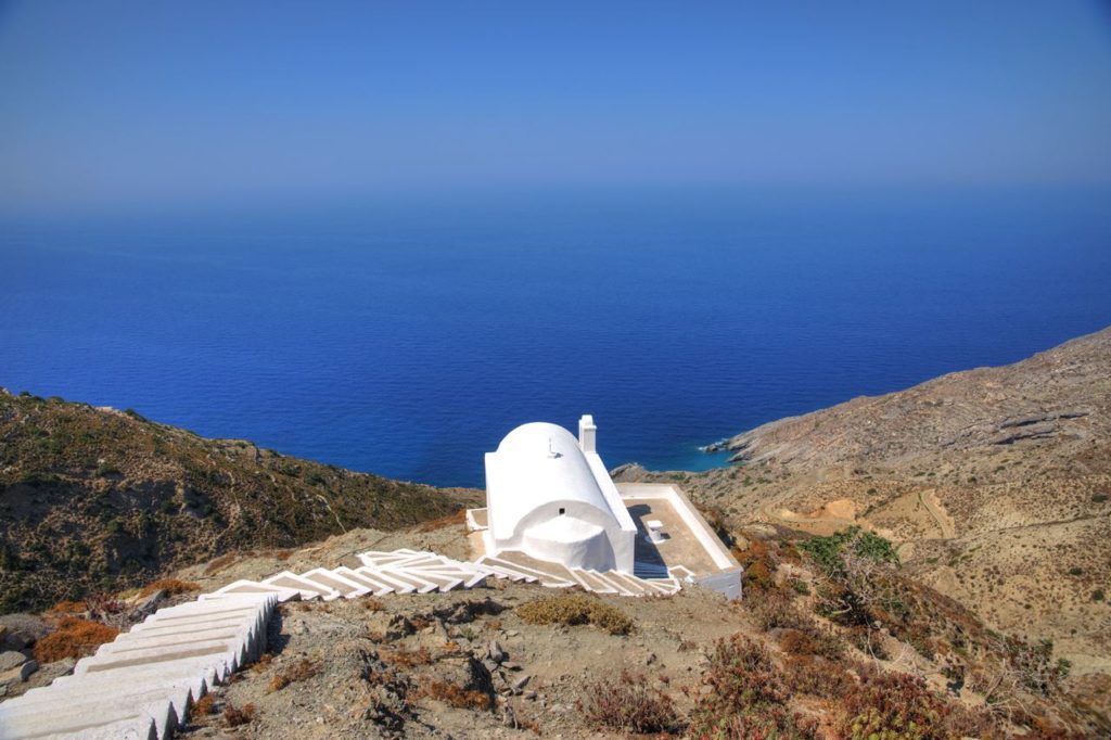 White domed chapel on a hillside overlooking the blue sea, with a winding white staircase down the slope.