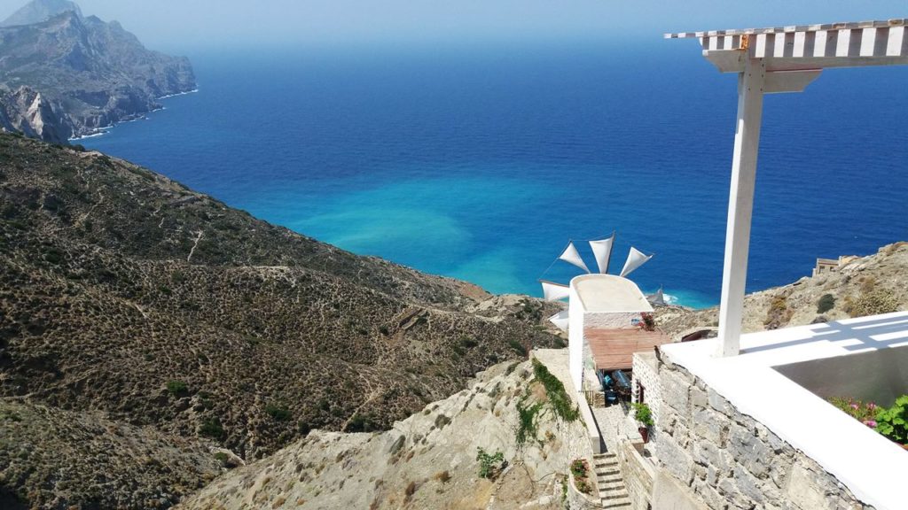 Whitewashed terrace and steps along a rugged cliff edge overlook a bright turquoise Aegean Sea.