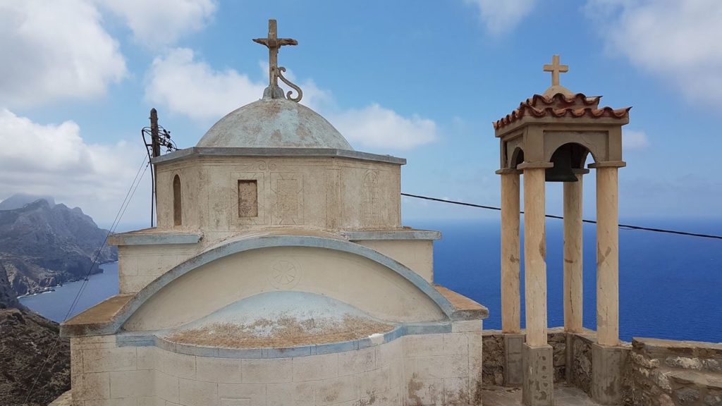 Small stone chapel with a dome and cross on top, overlooking the blue sea and cliffside scenery behind it.