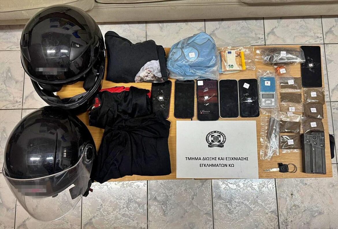 Evidence board: two black motorcycle helmets, assorted clothing, a blue bag, multiple smartphones, stacks of cash, and small bags likely containing drugs laid out on a table next to a Greek police sign.