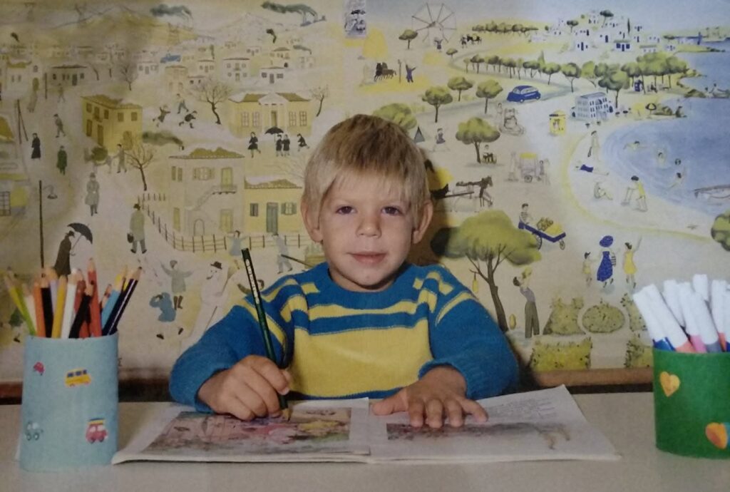 Young boy in a blue and yellow striped sweater sits at a table, drawing with a pencil amid cups of colored pencils and markers. The backdrop shows a whimsical mural wallpaper.