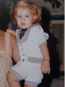 Young child with light hair wearing a white sailor romper with navy trim and gold buttons, standing with an adult's arm nearby.
