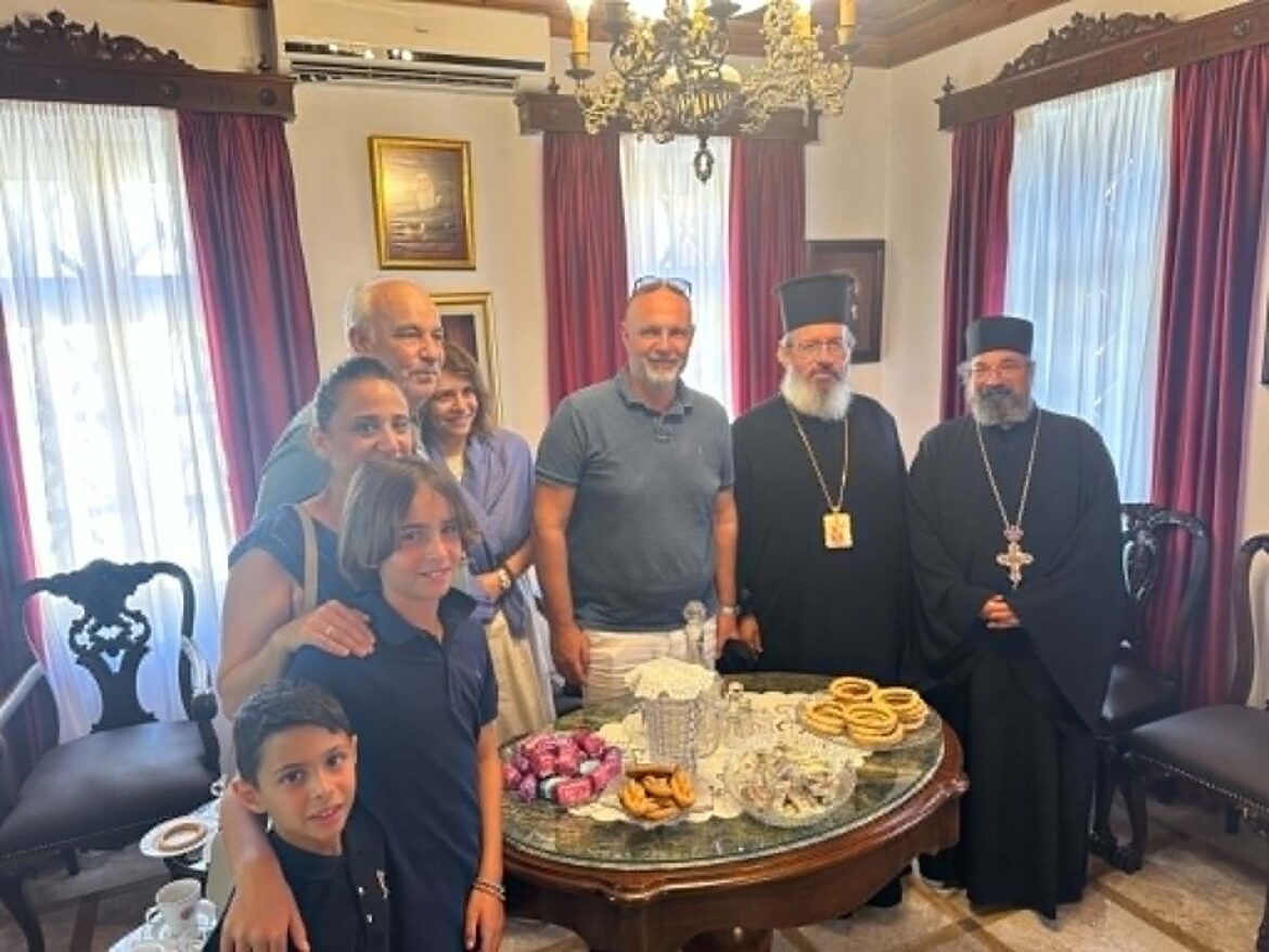Group photo around a cake and pastries on a table, with two Orthodox priests in black robes flanking guests in a dining room setting.