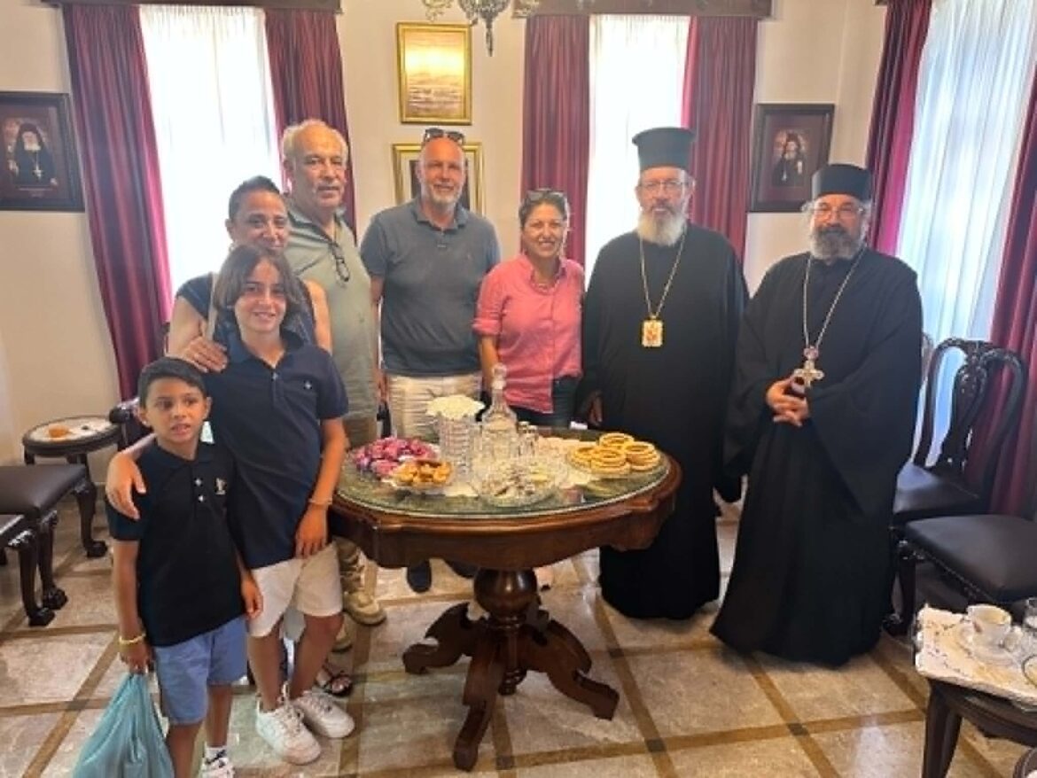 A diverse group of people posing around a round table with pastries and a cake, including two Orthodox clergy in black robes and hats, in a formal room with red curtains.