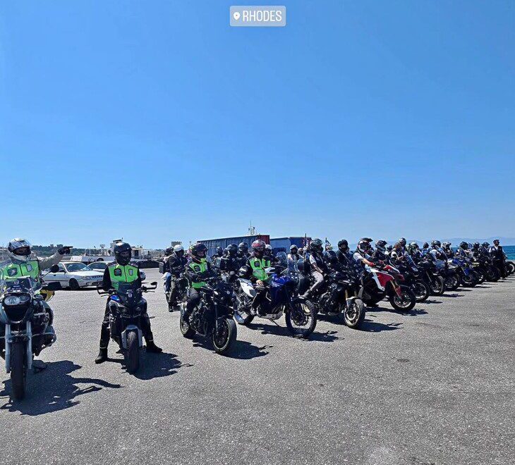 Line of motorcyclists wearing helmets and high-visibility jackets lined up in an open parking area by the coast near Rhodes